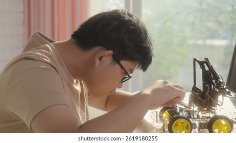 Close-up of Asian teenage boy connecting wires to circuit board of robotic car with robotic arm at home, concept of STEM education, robotics, and coding for young engineers.