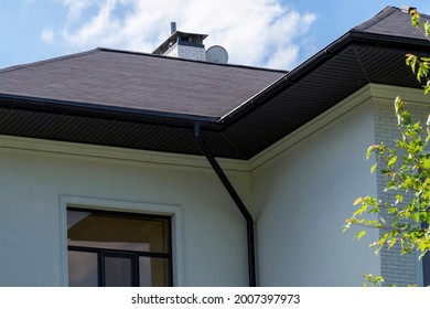 Close-up Of Asphalt Shingled Roofing Construction With Chimney, Plastic Soffit, Fascia Board And Roof Gutters Installed