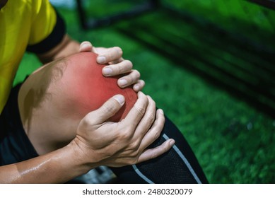 Close-up of an athlete holding a red, swollen knee, indicating injury or pain, with a blurred green field background. - Powered by Shutterstock