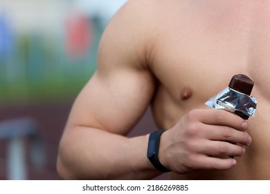 Close-up Of The Athletic Body Of A Man Holding An Energy Bar In His Hand In The Open Air. Healthy Eating, The Concept Of Outdoor Sports. A Muscular Athlete Eats A Protein Bar After A Workout