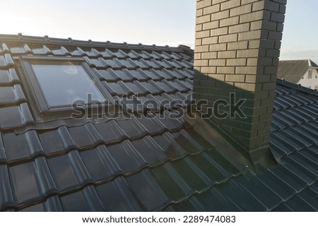 Closeup of attic window and brick chimney on house roof top covered with ceramic shingles. Tiled covering of building
