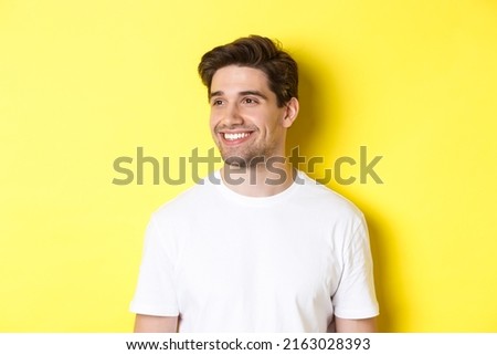 Close-up of attractive bearded man in white t-shirt smiling, looking left at copy space, standing against yellow background
