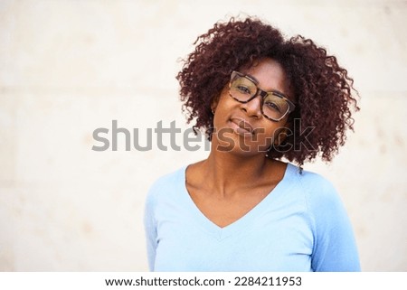 Close-up of attractive curly-haired woman wearing glasses, smiling pleasantly in a relaxing mood and posing with head tilted looking at camera, white wall in background and copy space. 
