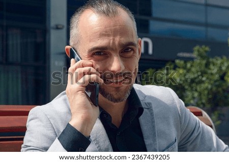 Close-up of attractive French Caucasian businessman wearing elegant dress clothes, on call talking on phone smiling and looking at camera sitting outdoors.