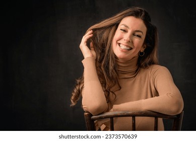 Close-up of an attractive middle aged woman with toothy smile wearing turtleneck sweater and sitting at isolated dark background. Copy space. Studio shot. Hand on forehead.