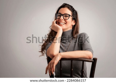 Close-up of an attractive middle aged woman with toothy smile wearing turtleneck sweater and eyewear while sitting at isolated grey background. Copy space. Studio shot. Hand on head.