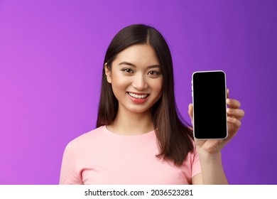 Close-up attractive pleasant female shop assistant present new phone app, hold smartphone show telephone screen, introduce application on display, stand purple background delighted