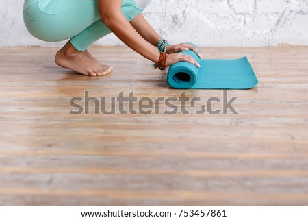 Close-up of attractive young woman folding blue yoga or fitness mat after working out at home in living room. Yoga and healthy lifestyle concept. Horizontal shot. white loft studio