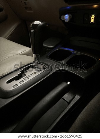 Close-up of an automatic car gear shift lever with visible transmission options and handbrake, taken inside a vehicle at night with soft interior lighting.