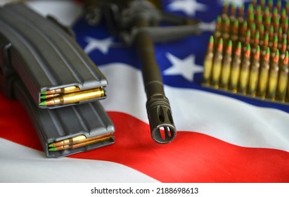 A closeup of an automatic carbine rifle's barrel over the US flag with stacks of ammunition rounds, 5.56 mm caliber. A concept image for gun control and the US military power. 