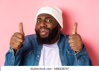 Close-up Of Awkward Black Man In Beanie Showing Thumbs-up But Feeling Cringe, Standing Unsure And Worried Against Pink Background