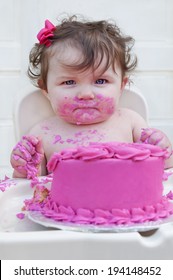 Closeup of a baby girl eating her first birthday smash cake with bright pink frosting looking into camera./Baby girl eating first birthday cake with pink frosting and bow in her hair