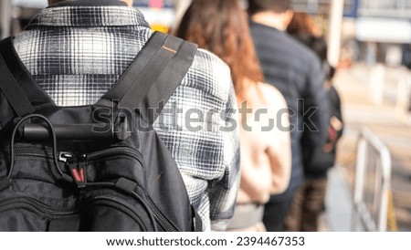 Close-up at back of a man with backpack bag during queue up and waiting to buy a attractive event ticket, with blurred background of many people in queue.