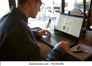 Close-up Back View Of Caucasian Businessman Hands Typing On Laptop Keyboard And Using Touchpad. Notebook And Pen On Foreground Of Workspace. Charts And Diagrams On Screen. Isolated No Face View.