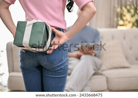 A close-up, back view image of a lovely wife holding and hiding a surprise gift present box behind her from her husband while he sits on a couch in the living room. anniversary, birthday gift