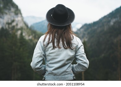 Closeup Back View Of Woman In Denim Jacket And Black Hat Looking At Mountain View, Isolated Dark Background. Rear View Of Female With Brown Hair Enjoying Peaceful Forest View