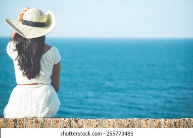 Closeup back view of woman sitting in white dress and hat looking out towards blue ocean and sky, isolated sea background