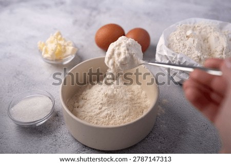 close-up of baking prep on a neutral countertop: a spoon pouring flour into a ceramic bowl with eggs, butter, sugar and an open paper bag of flour in a soft, warm studio kitchen scene, softlight