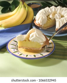 Close-up Of Banana Cream Pie Slice With Whole Pie, Coffee Cup, Leaves,and Bananas In Background.
