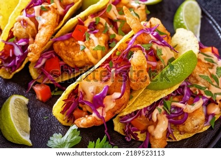 close-up of Bang Bang Shrimp Tacos with purple cabbage, tomatoes, parsley, lime drizzled with mayonnaise hot chili sauce on black plate on concrete table, horizontal view from above