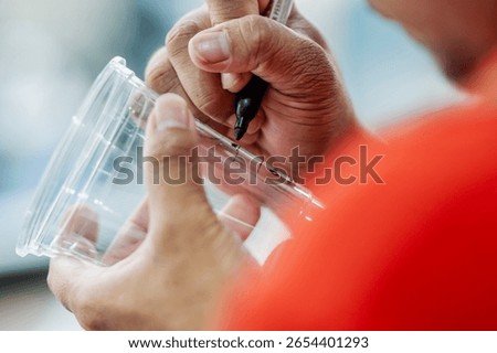 Close-up of a barista using a marker to write a customer's name on a clear plastic coffee cup for order identification.