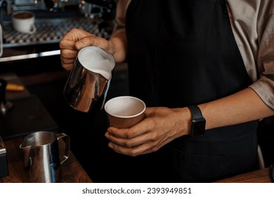 Close-up of a barista's hands pouring frothed milk into a paper coffee cup, with a blurred espresso machine in the background - Powered by Shutterstock