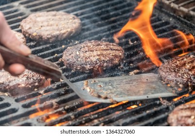 Closeup Of BBQ Burgers Patties On Hot Flaming Charcoal Grill. Top View Of Cookout Food Being Grilled With Flame And Utensil Flipping Burger.