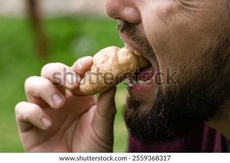 Close-up of a bearded man taking a bite of a small, flaky pastry in an outdoor setting with a green background.