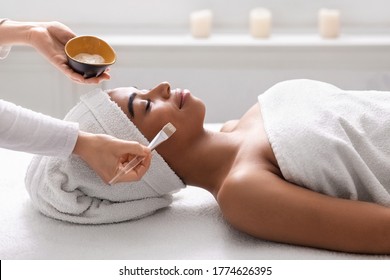 Closeup Of Beautician Holding Bowl With Face Mask Next To Sleeping Black Lady At Spa Salon