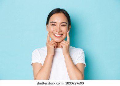 Close-up Of Beautiful Asian Girl Looking Pleased, Smiling White Teeth, Touching Cheeks, Recommend Skincare Product, Cosmetics For Facial Skin, Standing Light Blue Background