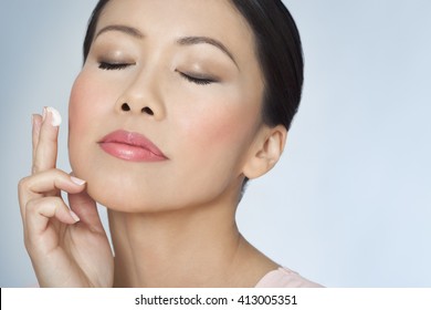 Closeup of beautiful Asian woman with glowing skin over thirty applying facial skincare cream with eyes closed. 