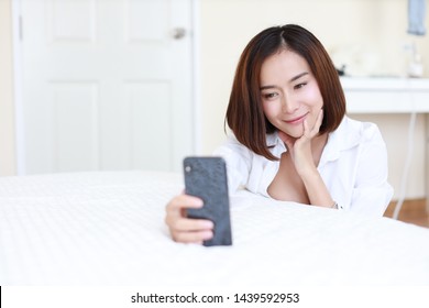 Closeup Beautiful Asian Woman, Short Hair In White Shirt Using Video Call Or Self Portrait Selfie From Mobile Phone On White Bed In Bedroom With Happy Smiling Face