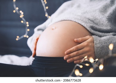 Close-up Beautiful Belly Of Pregnant Woman In Knitted Sweater In Light Of Garlands. Loving Mother Waiting A Baby. Concept Of Pregnancy, Maternity, Baby Shower Party, Holiday Winter Mood.