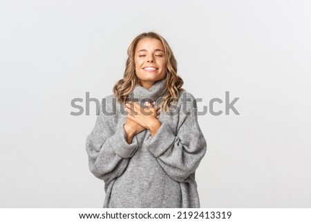 Close-up of beautiful blond girl, wearing grey sweater, feeling nostalgic, holding hands on heart and close eyes, daydreaming over white background