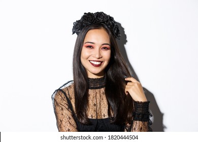 Close-up of beautiful elegant asian woman in black wreath and gothic lace dress smiling, standing over white background, dressed-up for halloween party