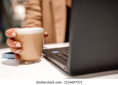 Close-up of beautiful female hands holding a large white cup of cappuccino. A business woman sits in a cafe working at a computer.