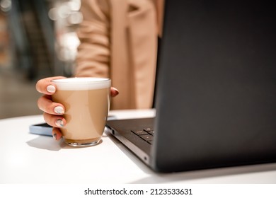 Close-up of beautiful female hands holding a large white cup of cappuccino. A business woman sits in a cafe working at a computer