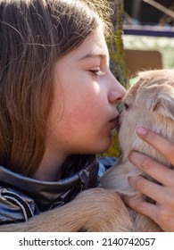 Close-up Of A Beautiful Girl Kissing A Little Goat, Real Life. Portrait Of A Teenage Girl Gently Hugging A Newborn Beige Baby Goat On The Street In The Sunlight. A Girl With A Goat.