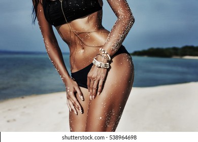 Close-up beautiful luxury slim girl in a black bikini on the beach the ocean. Sexy tanned body, flat stomach, perfect figure. Rest on a tropical island. Gold accessories. Photo in low key.