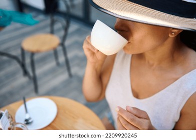 Close-up of a beautiful mysterious Latin American woman, in a straw hat covering half of her face, and elegant summer dress, sitting at a table in a summer terrace, enjoying her coffee break outdoors