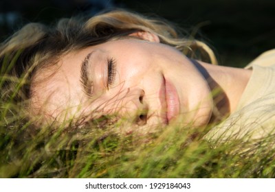 close-up of a beautiful teenage girl lying on green grass over sunlight. Eyes closed, enjoying nature outdoors. Cute teenage girl relaxing daydreaming in the park. Spring, summer, sunbathing