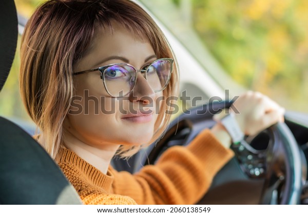 Close-up of a beautiful woman in\
glasses in a cozy brown sweater posing in the car holding a\
steering wheel blur. Concept training driving car instructor driver\
car 