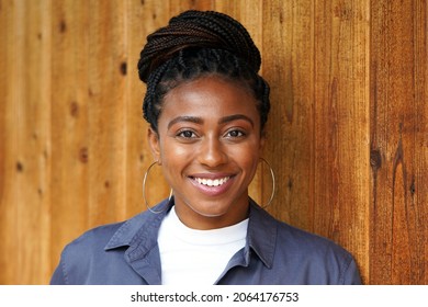 Closeup of beautiful young black professional woman wearing a braided bun hairstyle, white shirt and blue collared shirt stands against a wooden wall                             
