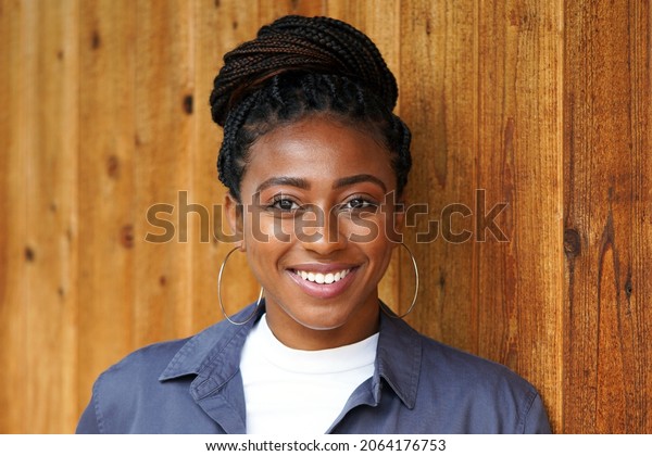 Closeup of\
beautiful young black professional woman wearing a braided bun\
hairstyle, white shirt and blue collared shirt stands against a\
wooden wall                            \
