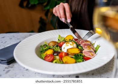 A close-up of a beautifully plated salad with seared tuna, fresh greens, cherry tomatoes, and a soft-boiled egg. A hand is using a knife and fork to enjoy the dish, with a glass of wine in background. - Powered by Shutterstock