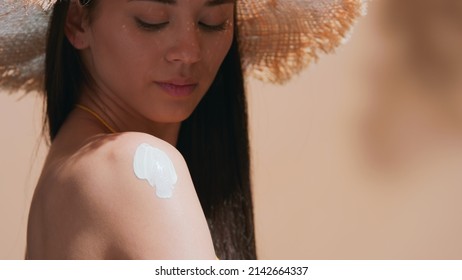 Close-up Beauty Portrait Of Dark-haired Girl In A Straw Hat Applying Sunscreen On Her Shoulder On Beige Background | Sunscreen Application Shot For Beauty Commercial