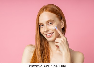 Closeup Beauty Portrait Of Redhead Caucasian Beautiful Female Applying Moisturizing Cream Or Face Primer And Looking At Camera Over Pink Background. Youth And Skin Care Concept.