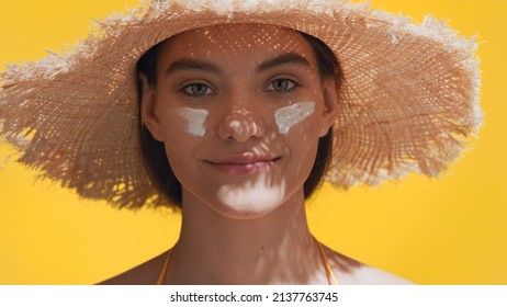 Close-up beauty portrait of young attractive dark-haired Caucasian woman in a straw hat having two smears of sunscreen cream on her face | Sunscreen application shot for face care commercial