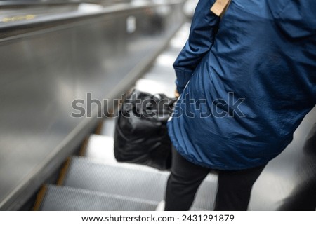 Close-up at behind of a person holding bag is standing on the escalator during moving down to the subway station. People in transportation activity. Selective focus.