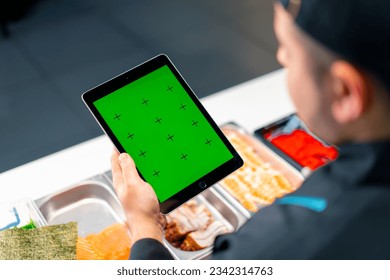 Close-up from behind the sushi chef holding a tablet with green screen open while making sushi and rolls - Powered by Shutterstock